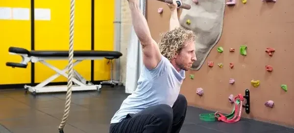 Man in a gym performing an overhead squat with a barbell in front of a climbing wall.