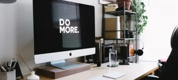 A modern, minimalist workspace featuring a large desktop computer on a wooden desk, with the screen displaying the motivational phrase “DO MORE.”
