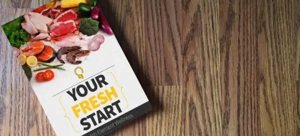 Booklet titled "Your Fresh Start" by 5th Element Wellness lying at an angle on a wooden floor, with a cover image of assorted fresh foods including meat, vegetables, and citrus.