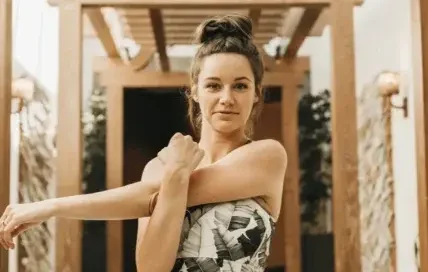 A woman is standing indoors under a wooden pergola-style structure, stretching one arm across her chest as if warming up or cooling down. She is wearing a matching patterned crop top and high-waisted skirt, and looking directly at the camera with a relaxed, confident expression.