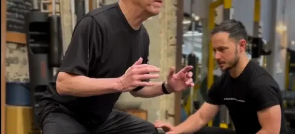 An older man is performing a forward lunge exercise in a gym while a younger trainer kneels nearby, closely observing and guiding his form.