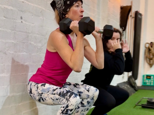 Over-40s personal training at 5th Element Wellness in Fitzroy North, with a coach mirroring correct movement while guiding a woman through a controlled dumbbell squat.