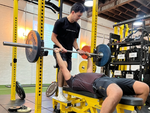 Returning to exercise after a long break at 5th Element Wellness in Fitzroy North, showing a coach safely spotting a client during a controlled barbell bench press.