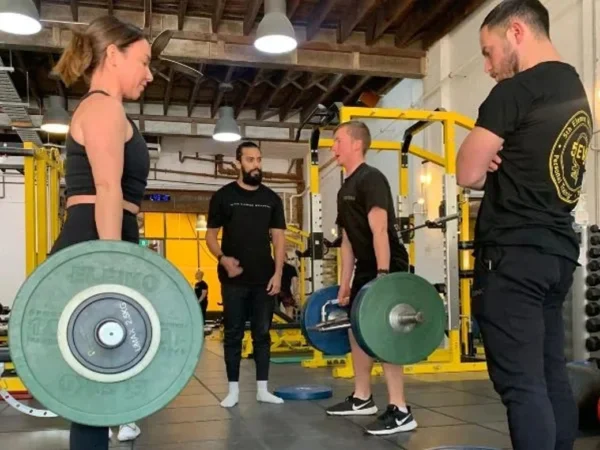 Woman holding barbell with weight plates during strength training session with three coaches in gym