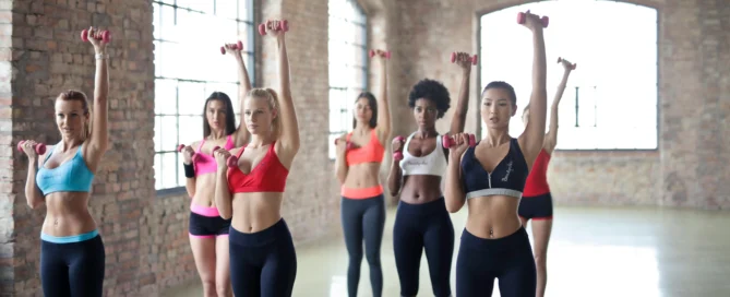 Six women in colorful athletic wear performing dumbbell exercises in an industrial-style gym with exposed brick walls and large arched windows